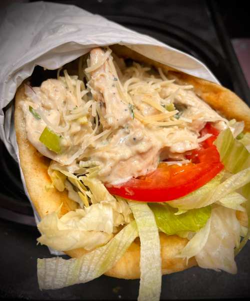 A pita filled with shredded chicken, lettuce, tomato, and a creamy sauce, wrapped in paper.