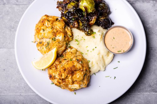 Two crab cakes with a lemon wedge, creamy mashed potatoes, and roasted Brussels sprouts on a white plate.