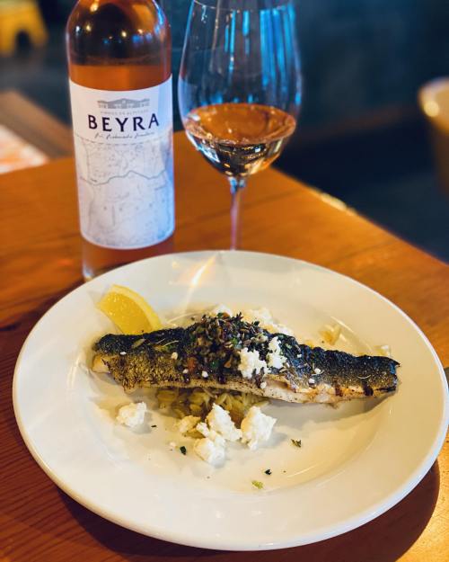 A plate of grilled fish with lemon and herbs, served with pasta, alongside a glass of rosé wine and a bottle.