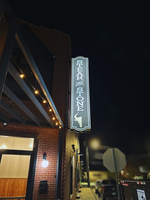 Sign for "Steak and Stone" illuminated at night, attached to a brick building with outdoor lighting.