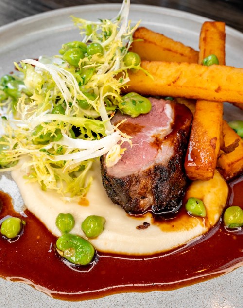 A plate featuring sliced meat, crispy fries, greens, and a creamy sauce, garnished with peas.