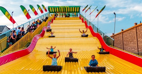 A colorful giant slide with children riding down on mats, surrounded by spectators and flags under a blue sky.