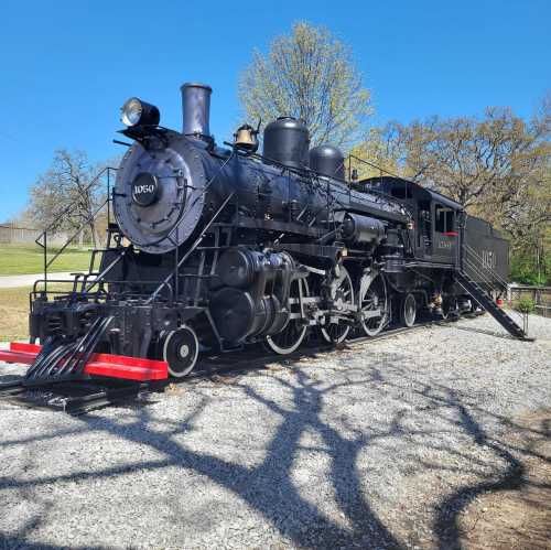 A vintage black steam locomotive displayed on a gravel path, surrounded by trees and a clear blue sky.