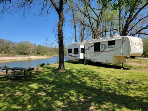 A parked RV by a river, surrounded by trees and grass, with a picnic table nearby under a clear blue sky.