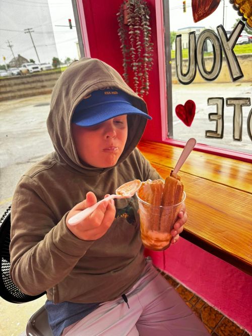 A child in a hoodie enjoys a dessert with a spoon, sitting by a window with a pink wall and greenery.
