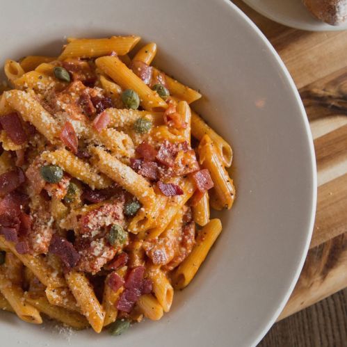 A bowl of penne pasta in a creamy sauce, topped with bacon, capers, and grated cheese, served on a wooden table.