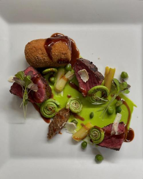 A gourmet dish featuring sliced beef, a croquette, green sauce, and garnishes on a white plate.