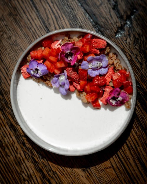 A plate of creamy dessert topped with strawberries, diced fruit, and edible flowers on a wooden table.