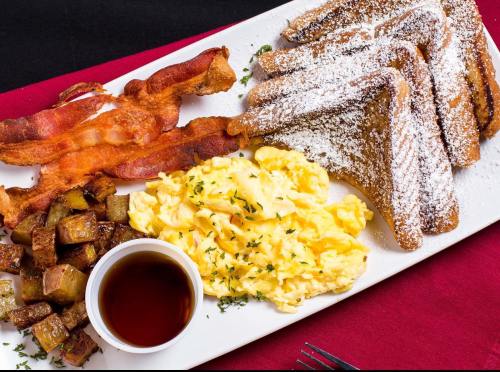 A breakfast plate featuring French toast, scrambled eggs, crispy bacon, and seasoned potatoes with syrup on the side.