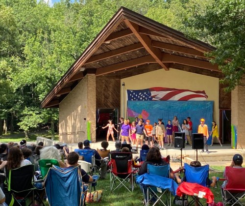A group of performers on stage in front of an audience seated on lawn chairs, with a large American flag backdrop.