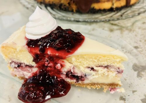 A slice of cheesecake topped with cherry sauce and whipped cream, with a whole chocolate-topped cake in the background.