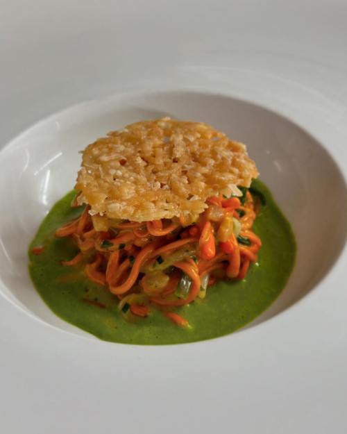 A colorful dish featuring spiralized vegetables topped with a crispy cheese wafer on a green sauce.
