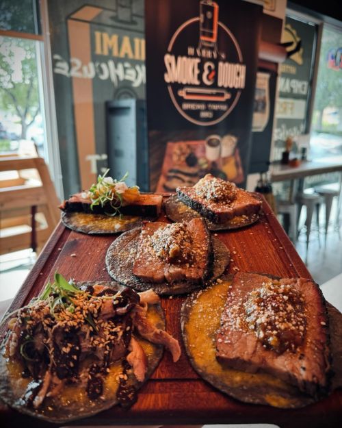 A wooden platter with five tacos topped with various meats and garnishes, set against a restaurant backdrop.