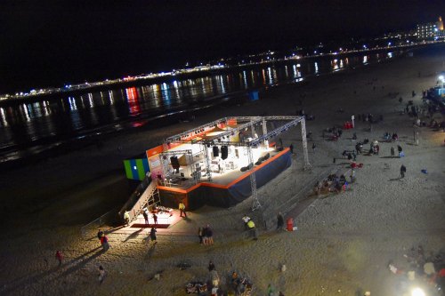 A beach at night with a stage setup, colorful lights, and people gathered on the sand.