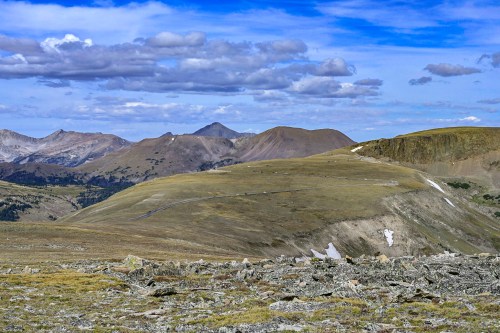 A panoramic view of rolling mountains under a blue sky with scattered clouds, showcasing grassy slopes and rocky terrain.