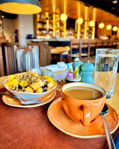 A bowl of fruit topped with granola beside a cup of coffee and a glass of water on a table in a cozy café.
