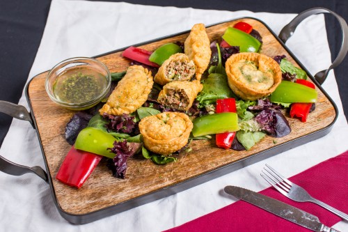 A wooden platter with assorted savory pastries on a bed of greens and bell peppers, accompanied by a dipping sauce.