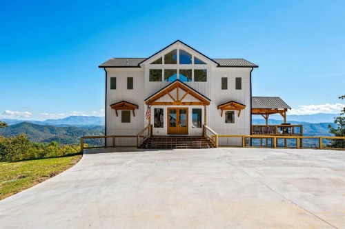 A modern mountain house with large windows, a wooden porch, and scenic views of rolling hills and blue skies.