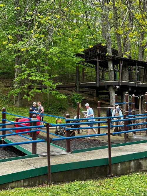 A group of people walking on a path in a park, with trees and a wooden structure in the background.