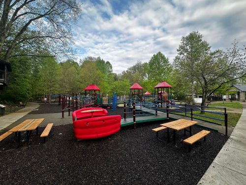 A colorful playground with red structures, picnic tables, and green trees under a cloudy sky.
