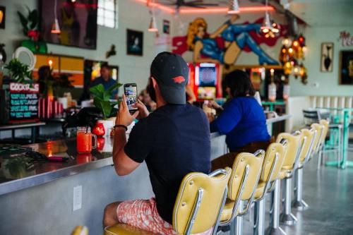 A person sits at a retro bar, taking a photo with their phone, while another patron enjoys a drink in the background.