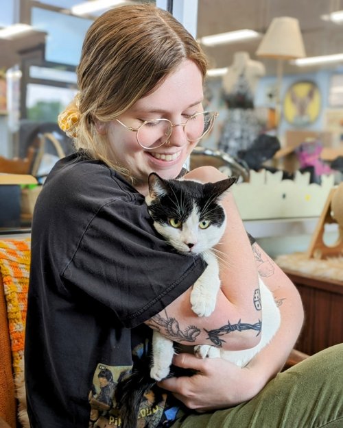 A smiling person with glasses hugs a black and white cat in a cozy, bright setting.