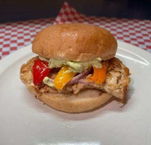 A grilled chicken sandwich with colorful peppers and sauce on a soft bun, served on a white plate.