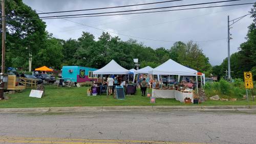 A vibrant outdoor market with tents, vendors, and colorful displays set against a green landscape and cloudy sky.