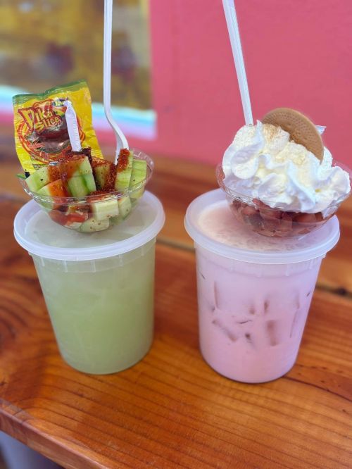 Two colorful drinks in clear cups: one green with fruit and a snack, the other pink topped with whipped cream and a cookie.