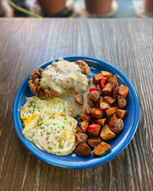 A blue plate with two fried eggs, crispy chicken with gravy, and seasoned roasted potatoes.