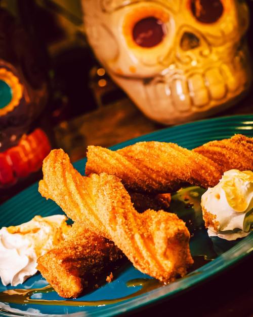 A plate of golden churros with whipped cream, set against colorful skull decorations.