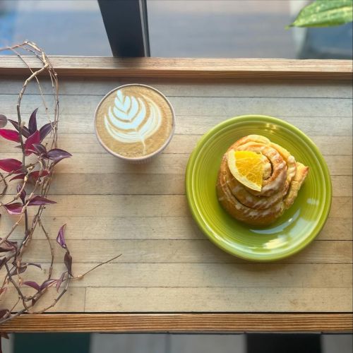A latte with leaf art beside a cinnamon roll topped with orange on a green plate, set on a wooden table.