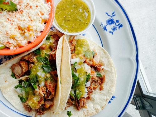 Two tacos filled with meat, onions, and cilantro, served with a side of green salsa and a bowl of elote.