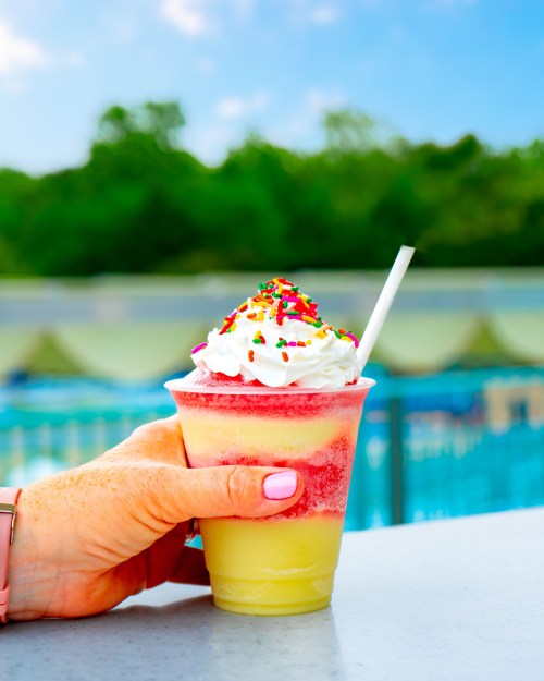 A hand holding a colorful layered drink topped with whipped cream and sprinkles, with a pool in the background.