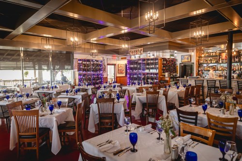 A spacious restaurant with tables set for dining, featuring blue glassware and a well-stocked bar in the background.