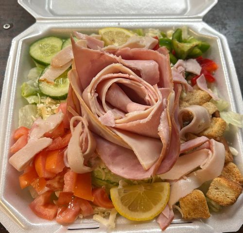 A salad topped with sliced ham arranged like a rose, surrounded by cucumbers, tomatoes, and croutons in a takeout container.