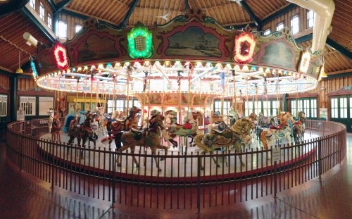 A colorful carousel with intricately painted horses, illuminated by bright lights, inside a wooden building.