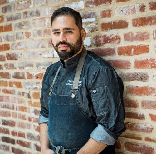A chef in a denim jacket and apron stands against a brick wall, looking confidently at the camera.