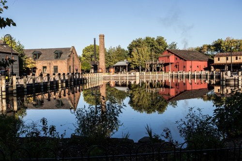 A serene waterfront scene with historic buildings and trees reflecting in calm water under a clear blue sky.