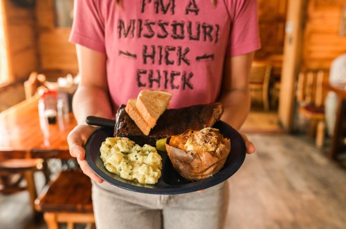 A person in a pink shirt holds a plate with ribs, baked potato, and potato salad in a rustic dining setting.