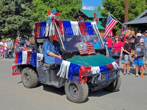 A decorated golf cart with red, white, and blue streamers and flags, driving in a parade with a crowd in the background.