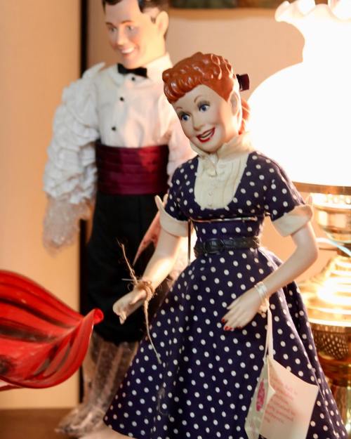 A vintage doll in a polka dot dress stands beside a tuxedoed figure, illuminated by a lamp in a cozy setting.