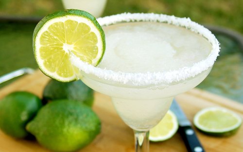 A refreshing margarita with a salted rim and lime garnish, surrounded by fresh limes on a wooden cutting board.