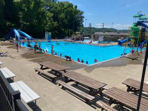 A sunny outdoor pool scene with children swimming, water slides, and picnic tables nearby.