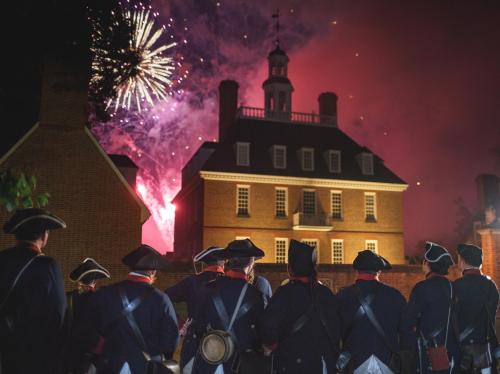 A group in historical attire watches fireworks illuminate a colonial-style building at night.