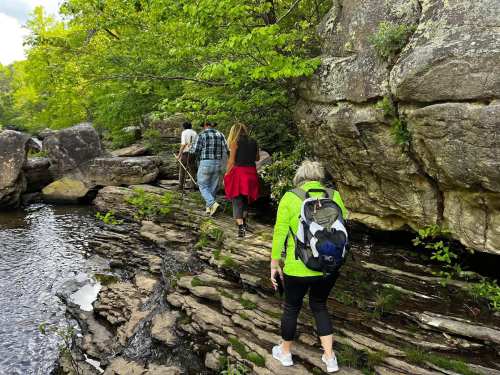 A group of hikers navigates rocky terrain near a stream, surrounded by lush greenery and large boulders.