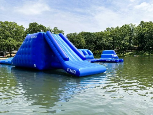 Two large blue inflatable water slides on a lake, surrounded by trees and a clear sky.
