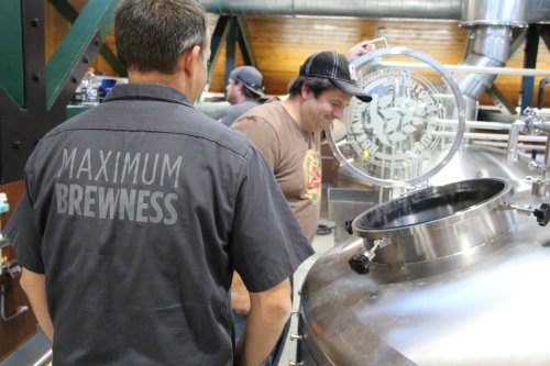 Two men in a brewery, one inspecting a brewing tank, the other wearing a shirt that says "Maximum Brewness."
