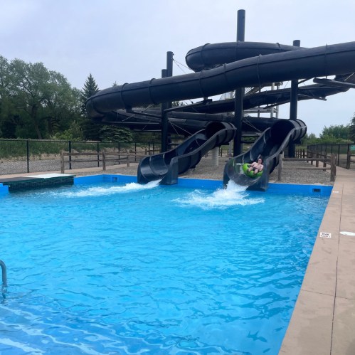 A water park scene featuring two water slides and a pool with splashing water. Trees are visible in the background.