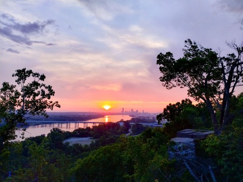 A vibrant sunset over a river, with city skyline in the distance and lush trees framing the scene.